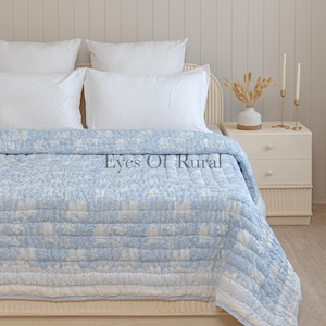 May include: A light blue and white floral patterned quilt on a bed with white pillows. The quilt has a striped border and the text "Eyes Of Rural". The bed is next to a white bedside table with candles and a vase.