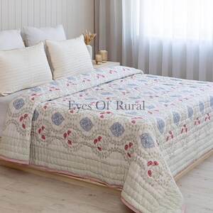 Könnte beinhalten: Eine weiße Steppdecke mit Blumenmuster und passenden Kissen auf einem Bett. Die Steppdecke hat ein sich wiederholendes Muster aus blauen und roten Blumendesigns auf weißem Hintergrund. Die Kanten sind mit einem rosa Rand versehen.