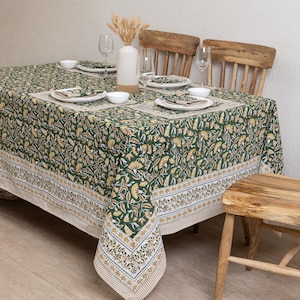 Puede incluir: Un mantel verde floral con manteles individuales y servilletas a juego. El mantel presenta un patrón repetido de flores amarillas y blancas sobre un fondo verde oscuro. La mesa está puesta con platos, cuencos y copas de vino blancos.