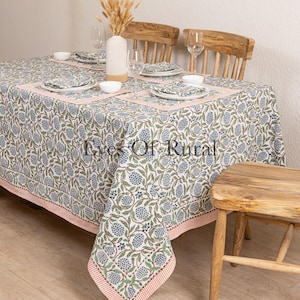 May include: A rectangular tablecloth featuring a floral design in blue, green, and white, with a pink border. The table is set with plates, napkins, and glasses. A vase with dried flowers is in the centre.