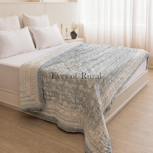 May include: A light blue and white patterned quilt with a quilted texture. The quilt is folded on a bed with white pillows and a white headboard. The text "Eyes of Rural" is visible on the quilt.