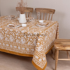Puede incluir: Un mantel amarillo mostaza con un patrón floral blanco y un borde blanco. El mantel está puesto sobre una mesa de madera con dos sillas de madera y un taburete de madera. El texto "Eyes of Rural" es visible en el centro del mantel.