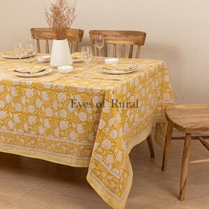 May include: A yellow tablecloth with a white floral pattern and a thin yellow border. The tablecloth is laid on a wooden table with two wooden chairs and a small wooden stool. The tablecloth is made of cotton and is perfect for a casual dining setting.