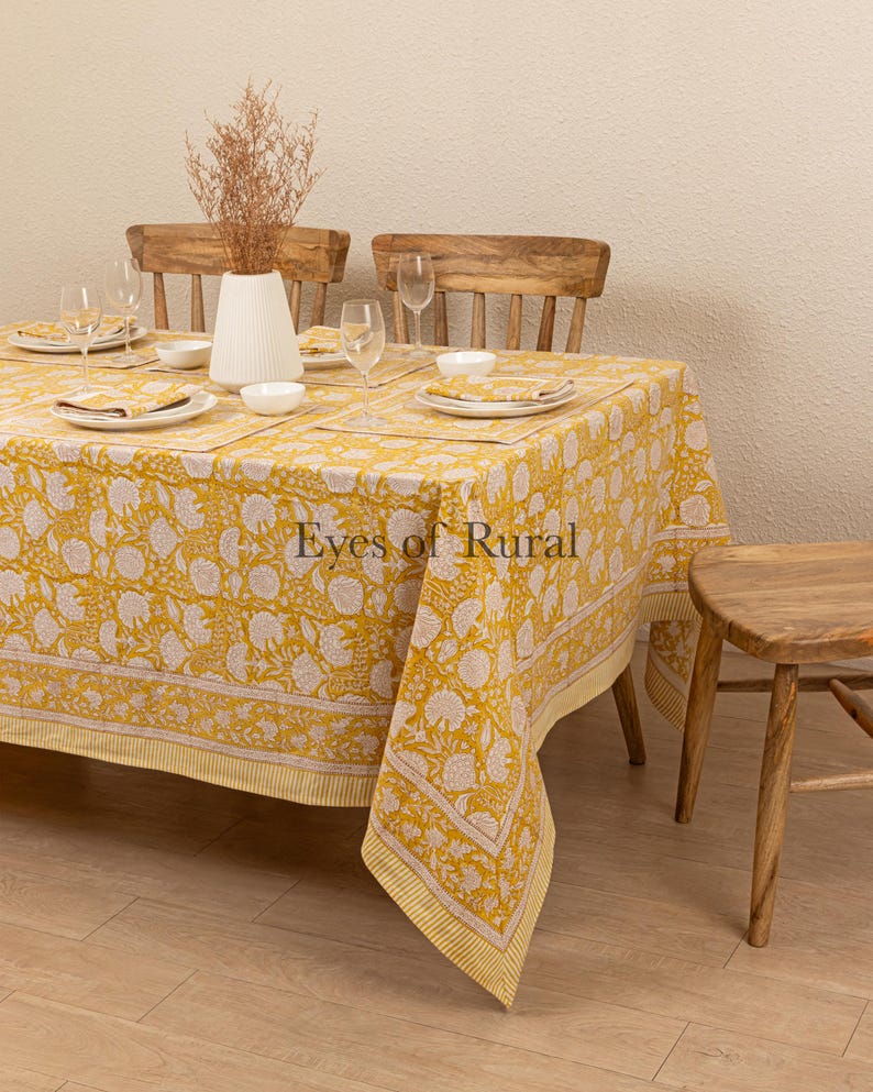 May include: A yellow tablecloth with a white floral pattern and a thin yellow border. The tablecloth is laid on a wooden table with two wooden chairs and a small wooden stool. The tablecloth is made of cotton and is perfect for a casual dining setting.