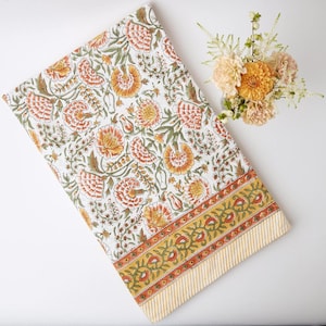 Puede incluir: Un mantel de mesa de algodón blanco con un estampado floral en tonos naranja y amarillo. El mantel tiene un borde amarillo y naranja con un patrón repetitivo.