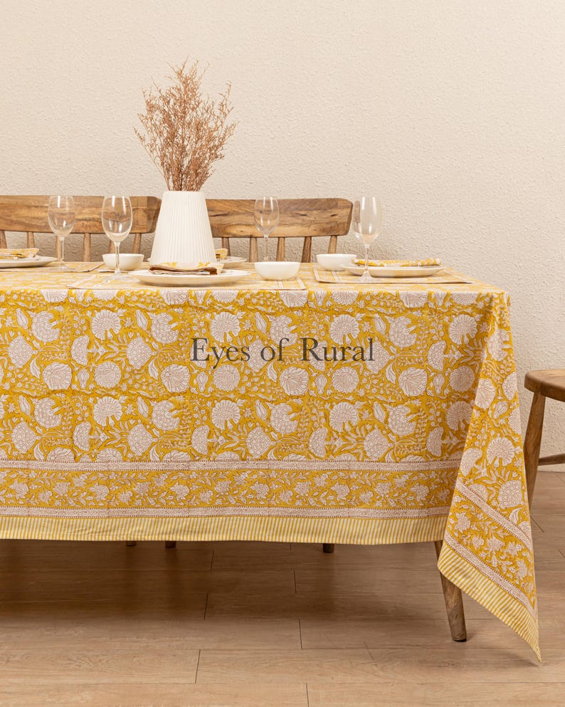 May include: A yellow tablecloth with a white floral pattern and a white border. The tablecloth is laid on a wooden table with four chairs and a vase of dried flowers in the center.