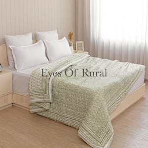 Op de afbeelding: Een bed met witte kussens en een groen-wit patroon quilt. De quilt heeft een geometrisch ontwerp en een rand met bladeren. De tekst "Eyes Of Rural" is zichtbaar op de quilt.