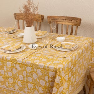 May include: A yellow tablecloth with a white floral pattern and a white border. The tablecloth is set with white plates, bowls, and napkins. There are two wooden chairs in the background.