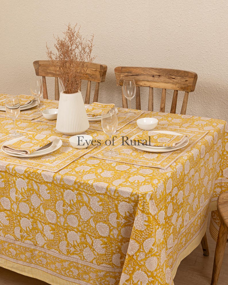May include: A yellow tablecloth with a white floral pattern and a white border. The tablecloth is set with white plates, bowls, and napkins. There are two wooden chairs in the background.
