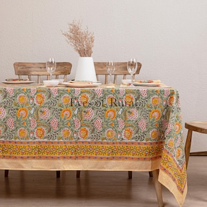 May include: A green and yellow floral tablecloth with a yellow border. The tablecloth is laid on a wooden table with four chairs and a vase of dried flowers.