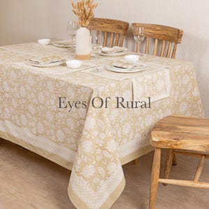May include: A beige floral tablecloth with matching placemats and napkins, set for a meal. The table is set with white plates, bowls, and wine glasses. The text "Eyes Of Rural" is visible on the tablecloth.