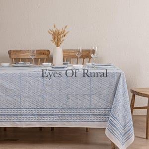 May include: A blue and white patterned tablecloth covers a wooden table set for a meal. The tablecloth has a repeating geometric design and a border with a leaf pattern. The text "Eyes Of Rural" is visible on the tablecloth.