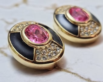 Puede incluir: Un par de pendientes ovalados dorados. Cada pendiente presenta una gran gema rosa ovalada en el centro, rodeada por un borde de esmalte negro y cristales transparentes. Los pendientes tienen un diseño clásico y elegante.