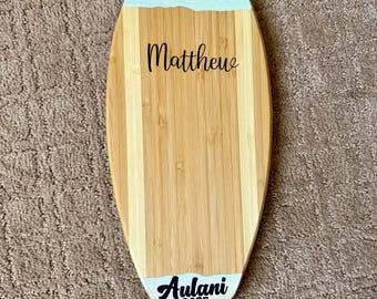 Può includere: Un tagliere a forma di tavola da surf in bambù con la parte superiore dipinta di blu e bianco. Il nome "Matthew" è scritto in corsivo nero. Il fondo riporta la scritta "Aulani 2025" in corsivo nero.