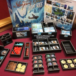 May include: A black and grey organiser for the board game "Sleeping Gods: Distant Skies". The organiser includes compartments for cards, tokens, and miniatures. The organiser is made of high-quality materials and is designed to fit perfectly in the game box.