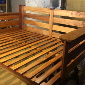 May include: A wooden bed frame with a slatted base. The frame is made of light brown wood with a darker brown stain. The bed frame is in a workshop setting.