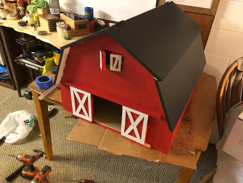 May include: A handcrafted red barn with a black roof and white accents. The barn features two white doors with an X design and a small window. This miniature barn is likely for decorative purposes or as a toy.