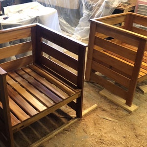 May include: Two wooden chairs with slatted backs and seats. The chairs are unfinished and have a natural wood color. They are sitting on a wooden floor in a workshop.
