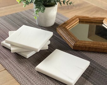 May include: Four white marble coasters with a subtle veining pattern, stacked on a brown and gray textured surface. A small potted plant with green leaves is in the background.