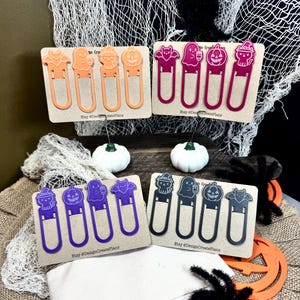 May include: Halloween-themed bookmarks in orange, pink, purple, and black, each featuring a different spooky character. The bookmarks are displayed on cardboard backing, with a white canvas pouch filled with pens and markers in the foreground.