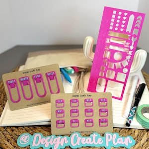 May include: A collection of planner accessories, including pink and gold stencils, paper clips, and month labels. The accessories are on a notebook with pens and washi tape. The text "Design Create Plan" is visible.