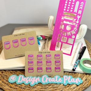 May include: A collection of planner accessories, including pink die-cut stencils, calendar tabs, and labels. The items are arranged on a notebook and a woven mat. The text "Design Create Plan" is visible on several items.