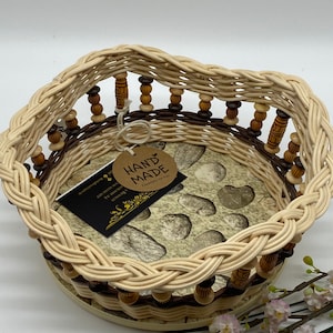 Könnte beinhalten: Ein geflochtener Weidenkorb mit braunem und beigefarbenem Muster. Der Korb hat einen runden Boden und einen dekorativen Rand mit Holzperlen. Der Korb ist mit einem kleinen Etikett "Hand Made" gekennzeichnet.