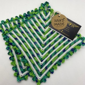 Könnte beinhalten: Zwei grüne, weiße und blaue gehäkelte Topflappen mit einem gewellten Rand. Die Topflappen sind übereinander gestapelt. Ein braunes Etikett mit dem Text "HAND MADE" ist am oberen Topflappen befestigt.
