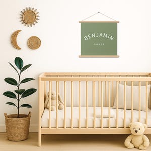May include: A nursery scene with a wooden crib, a green wall hanging with the name "BENJAMIN PARKER", and decorative wall art. A stuffed bunny and teddy bear sit in the crib. A potted plant and woven sun and moon decor complete the room.