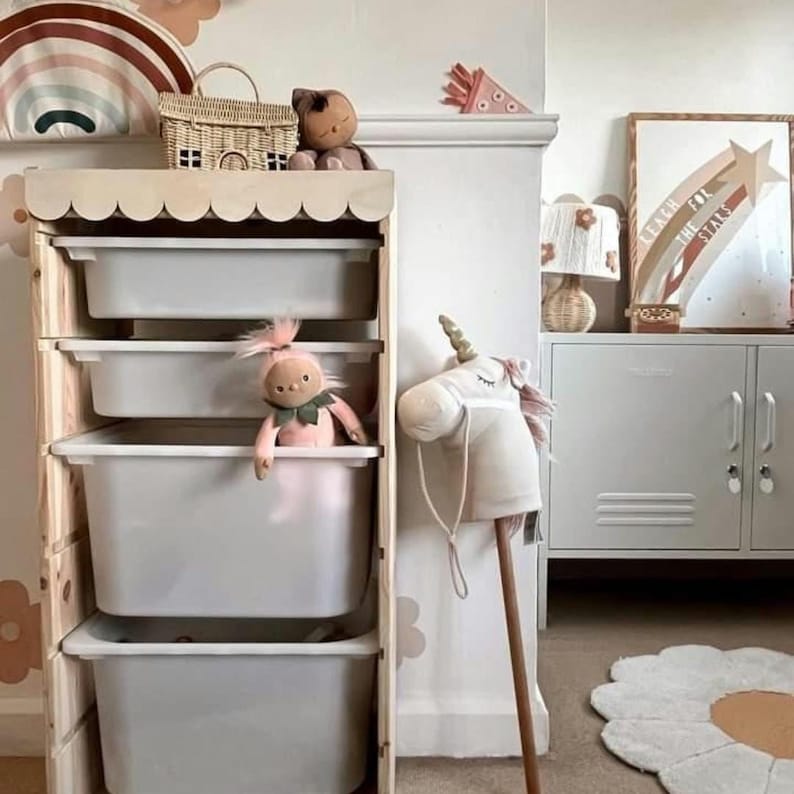 May include: A wooden storage unit with four white plastic drawers. The unit has a scalloped top edge and is decorated with rainbow and flower decals. A pink doll, a stuffed unicorn, and a wicker basket are on top of the unit.