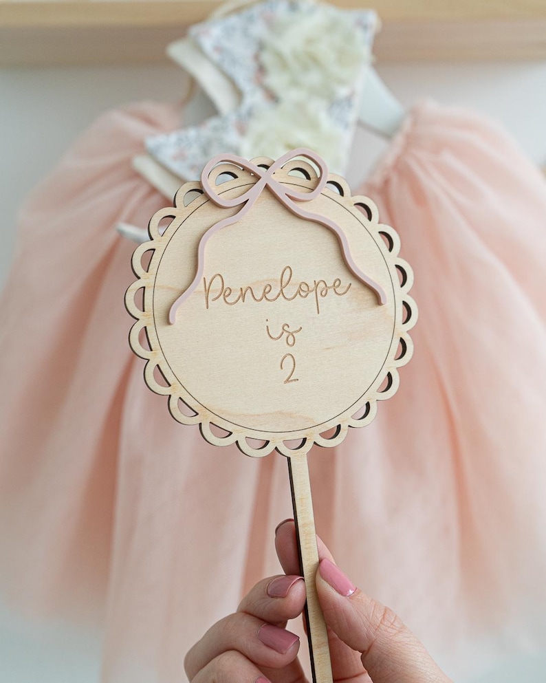 May include: A wooden sign with a pink bow and the text "Penelope is 2" on a stick. The sign is held by a hand.