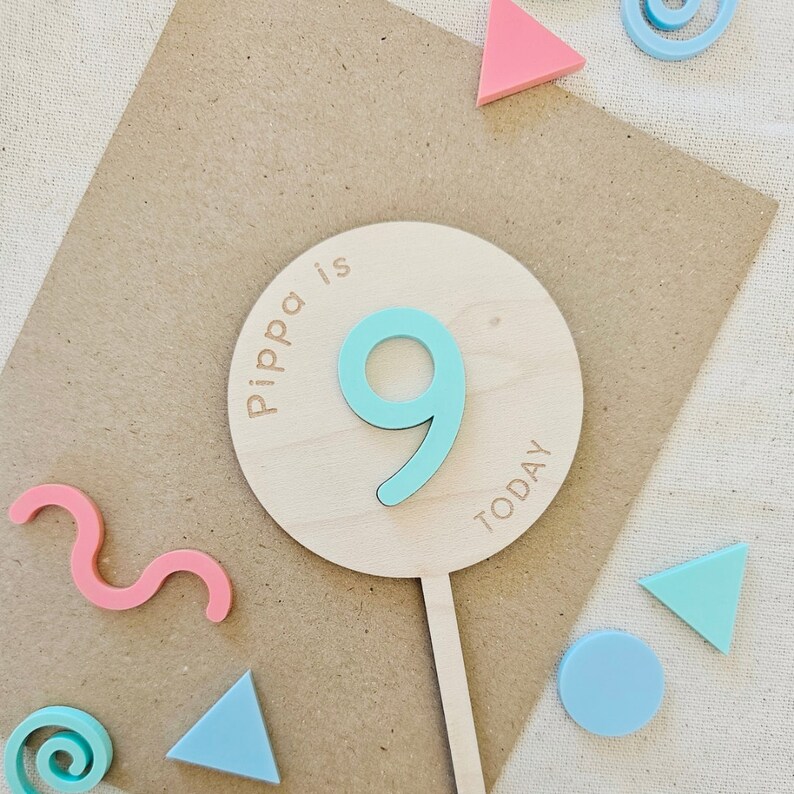 May include: A wooden cake topper with the text "Pippa is 9 today" in a circle. The number 9 is a light blue color and the text is brown. The cake topper is on a brown background with colorful geometric shapes.