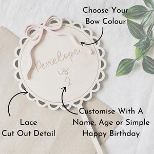 May include: A wooden cake topper with a lace cut-out design. The topper features a pink bow and the text "Penelope is 2".