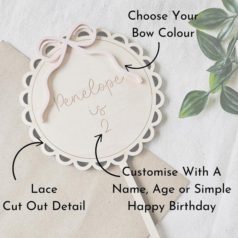 May include: A wooden cake topper with a lace cut-out design. The topper features a pink bow and the text "Penelope is 2".
