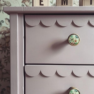 May include: A light purple dresser with scalloped drawer edges and floral print knobs. The dresser has two drawers visible, with a decorative edge. The knobs have a gold trim and a floral design.