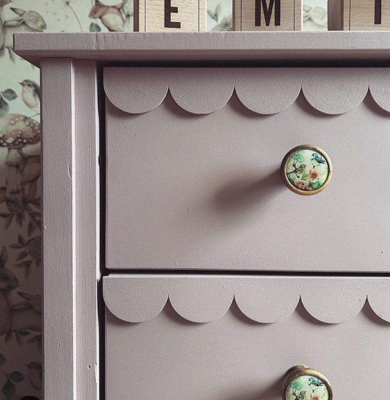 May include: A light purple dresser with scalloped drawer edges and floral print knobs. The dresser has two drawers visible, with a decorative edge. The knobs have a gold trim and a floral design.