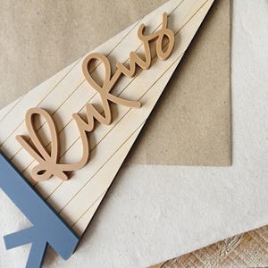 May include: A wooden pennant with the word "Kws" in a cursive font. The pennant is light brown with horizontal lines, and the base is a blue-gray color. The background includes a tan paper and a white fabric.