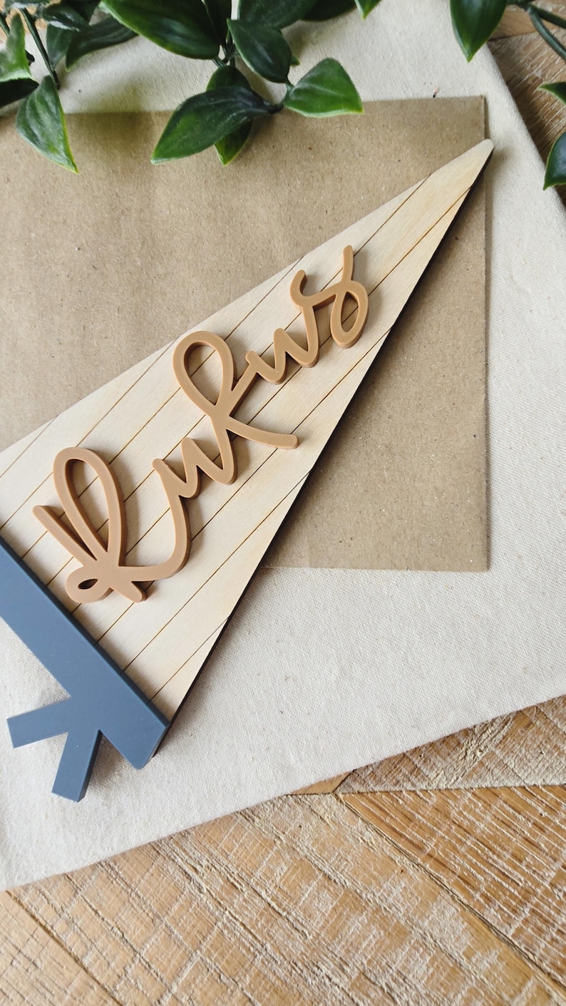 May include: A wooden pennant with the word "Kws" in a cursive font. The pennant is light brown with horizontal lines, and the base is a blue-gray color. The background includes a tan paper and a white fabric.