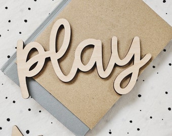 May include: Wooden sign with the word "play" written in a cursive font. The sign is on a brown background with a white and black polka dot pattern.