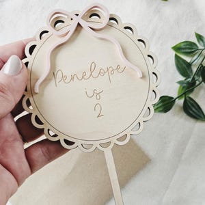 May include: A wooden cake topper with a pink bow and the text "Penelope is 2" in a decorative circle.