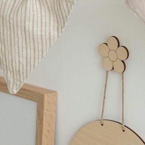 May include: A light-colored wooden wall hook shaped like a five-petaled flower with a dark brown edge. A thin, light brown string hangs from the hook, supporting a circular wooden plaque. A beige and white striped fabric hangs in the upper left corner.