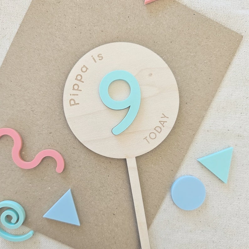 May include: A wooden cake topper with a light blue number 9 and the text "Pippa is Today" in a circle. The topper is on a brown background with pink and blue geometric shapes.