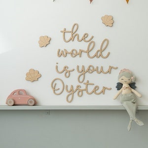 May include: Wooden wall decor featuring the phrase "The world is your oyster" in cursive script. There are also small wooden seashells and a mermaid doll on the wall.