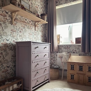 May include: A child's bedroom with a lavender dresser, wooden shelf, and a dollhouse. The dresser has five drawers with scalloped edges and brass knobs. The wallpaper features a whimsical pattern of mushrooms, birds, and flowers. A gray crocheted rug sits on the floor.