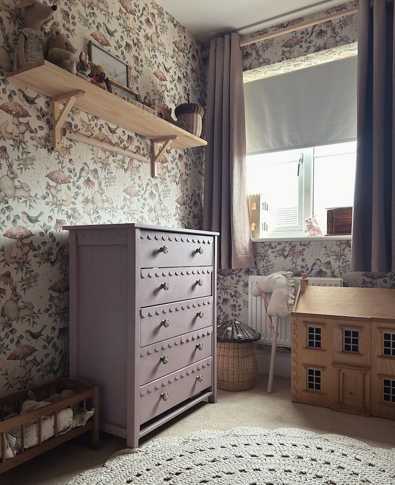 May include: A child's bedroom with a lavender dresser, wooden shelf, and a dollhouse. The dresser has five drawers with scalloped edges and brass knobs. The wallpaper features a whimsical pattern of mushrooms, birds, and flowers. A gray crocheted rug sits on the floor.