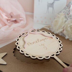 May include: A wooden wand with a pink ribbon and a circular wooden sign that says "Penelope is 2".