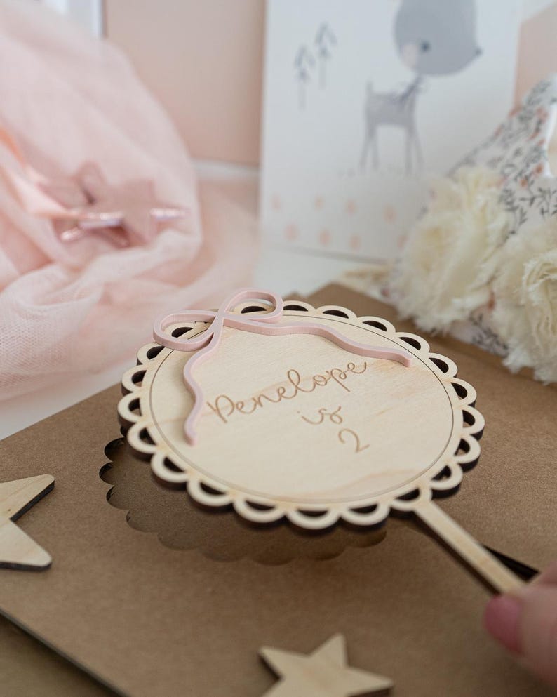 May include: A wooden wand with a pink ribbon and a circular wooden sign that says "Penelope is 2".