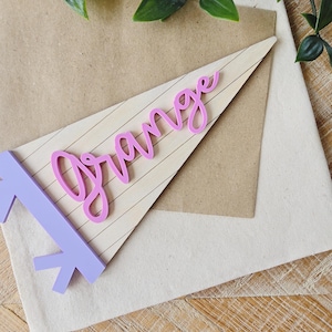 May include: A light purple pennant with the word "Orange" in pink script. The pennant is on a light brown background with a white fabric and a wooden surface. Green leaves are in the background.