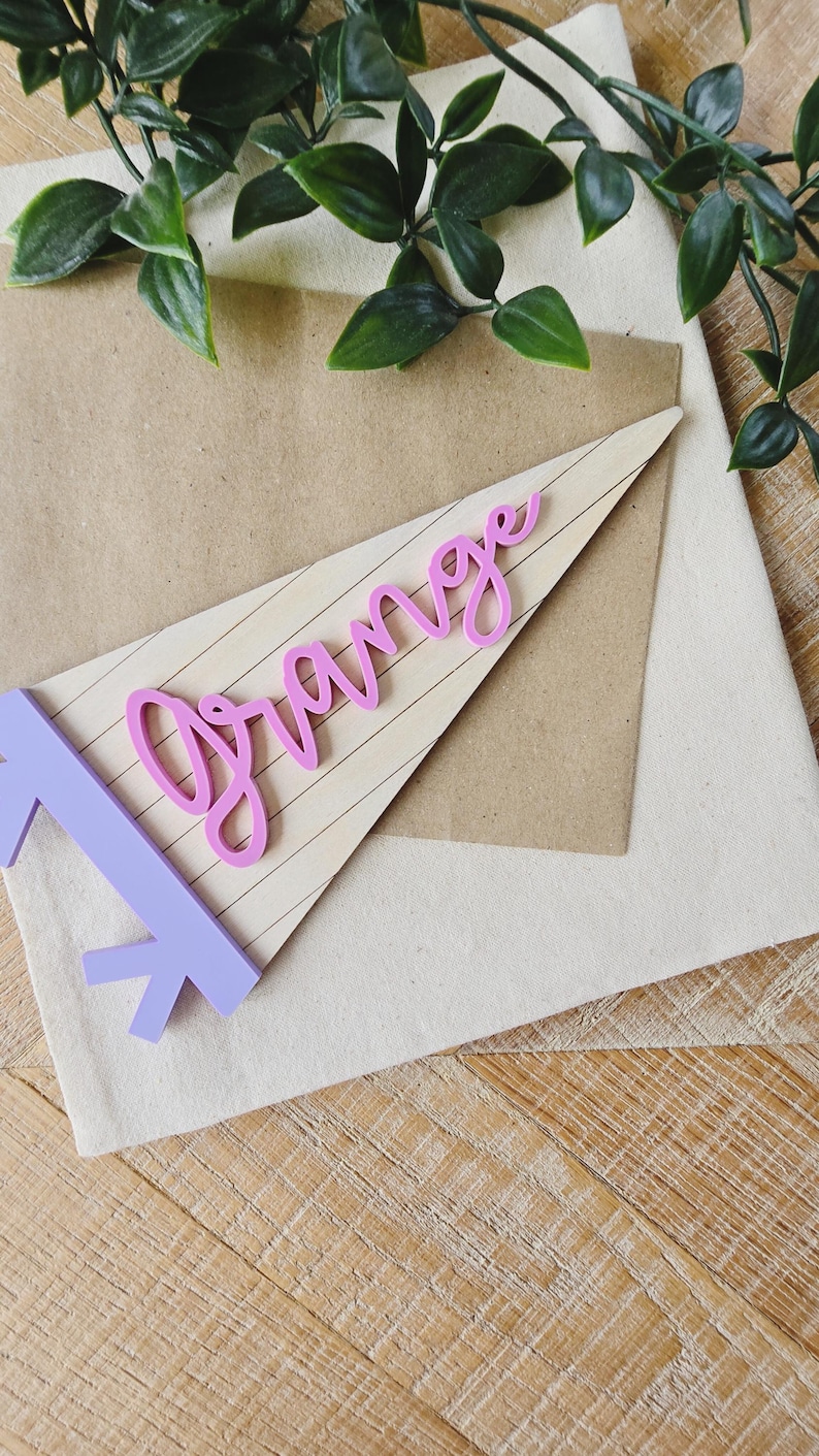 May include: A light purple pennant with the word "Orange" in pink script. The pennant is on a light brown background with a white fabric and a wooden surface. Green leaves are in the background.