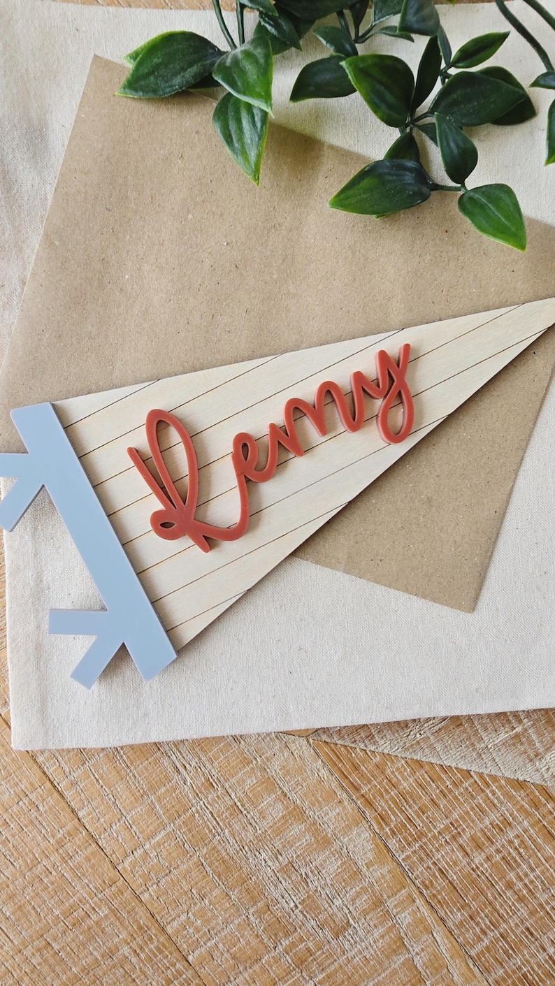 May include: A wooden pennant with the name "Remy" in red script. The pennant is light-colored with horizontal lines and a light blue arrow design on the left. The pennant rests on a brown envelope and a white cloth.